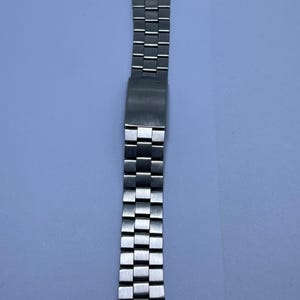 Puede incluir: Una correa de reloj plateada con un patrón de cuadros. La correa es de metal y tiene un cierre. La correa del reloj se muestra sobre un fondo azul claro.
