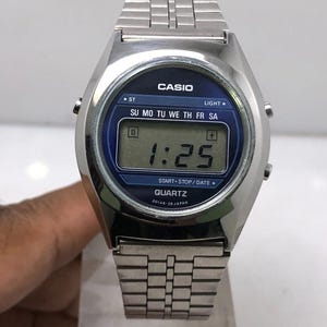 May include: A silver-toned Casio digital watch with a blue face displaying the time as 1:25. The watch has a rectangular digital display and a silver metal band. The watch face includes the text "CASIO" and "QUARTZ".