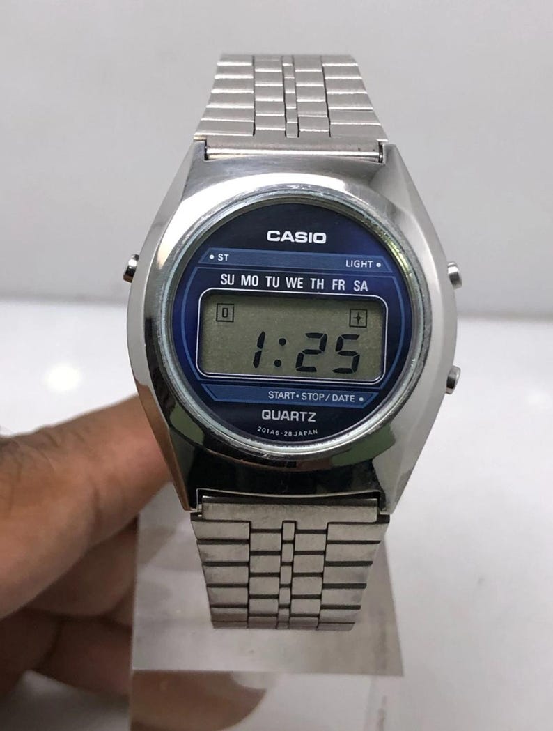 May include: A silver-toned Casio digital watch with a blue face displaying the time as 1:25. The watch has a rectangular digital display and a silver metal band. The watch face includes the text "CASIO" and "QUARTZ".