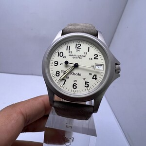 Może przedstawiać: Srebrny zegarek kwarcowy Hamilton Khaki z kremową tarczą, czarnymi cyframi i wskaz&oacute;wkami. Zegarek posiada datownik i szary sk&oacute;rzany pasek. Zegarek ma średnicę około 3,8 cm.