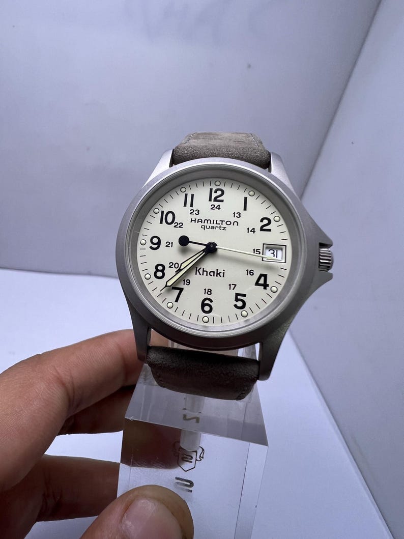 Może przedstawiać: Srebrny zegarek kwarcowy Hamilton Khaki z kremową tarczą, czarnymi cyframi i wskaz&oacute;wkami. Zegarek posiada datownik i szary sk&oacute;rzany pasek. Zegarek ma średnicę około 3,8 cm.