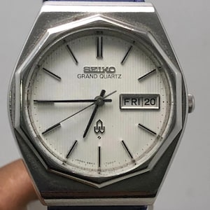 Puede incluir: Un reloj Seiko Grand Quartz plateado con esfera blanca y correa de cuero azul. La esfera muestra el día y la fecha, con "FRI 20" visible. El reloj tiene agujas y marcadores negros.