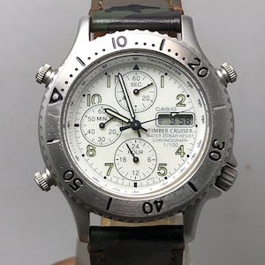 May include: A silver Casio Timber Cruiser chronograph watch with a white face, black hands, and multiple sub-dials. The watch features a digital display and a camouflage-patterned leather strap. The watch is water-resistant and has a tachymeter bezel.
