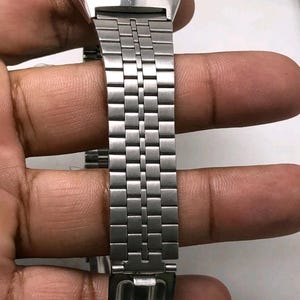 May include: A silver-toned digital watch with a blue display. The watch features a rectangular face and a stainless steel band with a repeating rectangular link pattern. The watch is held by a hand.