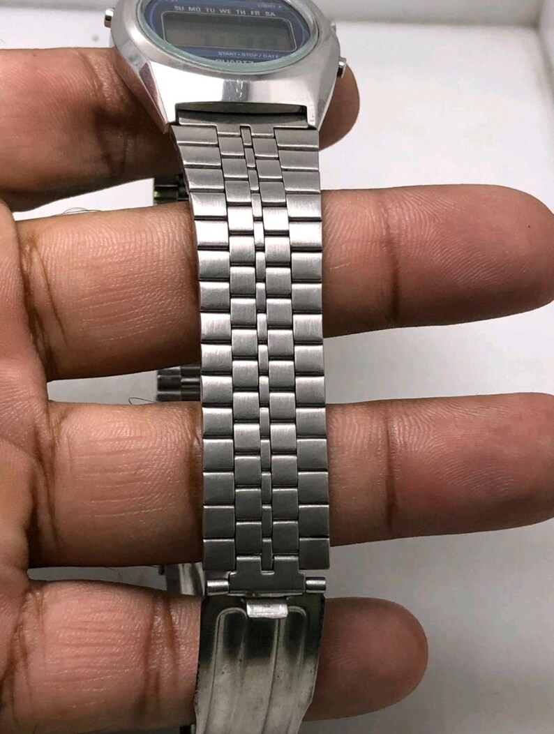 May include: A silver-toned digital watch with a blue display. The watch features a rectangular face and a stainless steel band with a repeating rectangular link pattern. The watch is held by a hand.