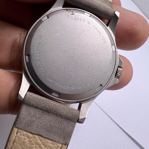 Może przedstawiać: Zbliżenie na stalowy zegarek ze sk&oacute;rzanym, szarym paskiem. Tarcza zegarka jest srebrna z napisem "HAMILTON" i "WATER RESISTANT 3 ATM". Pasek jest w jasnobrązowym kolorze.