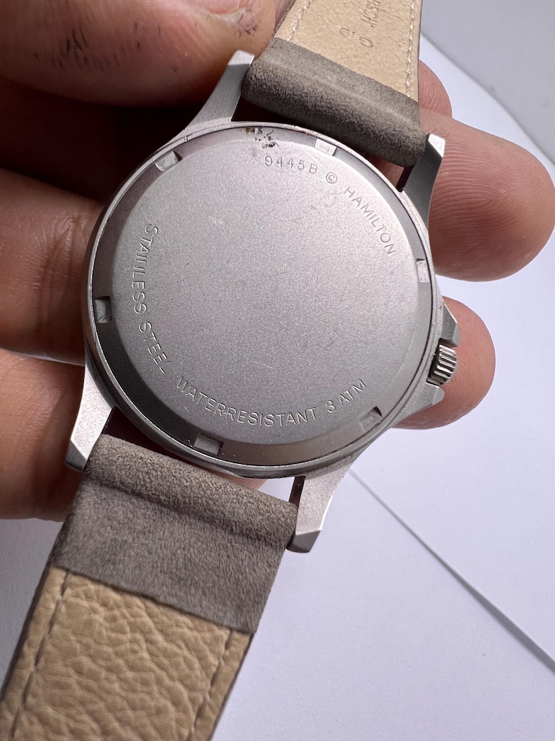 Może przedstawiać: Zbliżenie na stalowy zegarek ze sk&oacute;rzanym, szarym paskiem. Tarcza zegarka jest srebrna z napisem "HAMILTON" i "WATER RESISTANT 3 ATM". Pasek jest w jasnobrązowym kolorze.