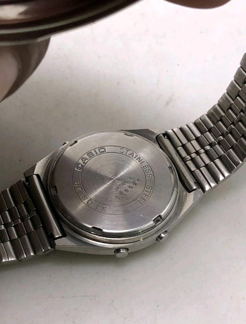 May include: A close-up of the back of a silver Casio watch with a stainless steel band. The watch face is silver with the words "CASIO STAINLESS STEEL JAPAN B 600595" engraved on it.