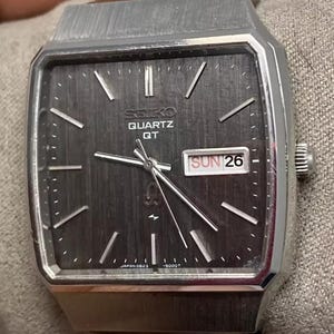 May include: A vintage Seiko Quartz QT watch with a dark gray face and silver hands. The watch has a rectangular case and a date display showing "SUN 26". The watch is a classic timepiece.