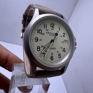 Może przedstawiać: Srebrny zegarek kwarcowy Hamilton Khaki z kremową tarczą, czarnymi wskaz&oacute;wkami i datownikiem. Zegarek ma jasnobrązowy sk&oacute;rzany pasek. Tarcza zegarka wyświetla cyfry od 1 do 12, z dodatkowymi cyframi 13-24.