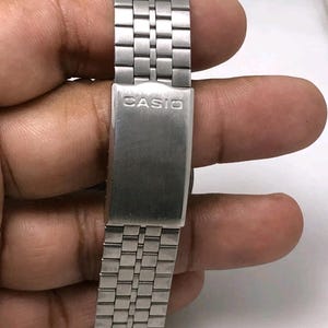 May include: Close-up of a silver-toned watch band with the brand name "CASIO" engraved on the clasp. The band is made of rectangular metal links. The watch band is held by a person's hand.
