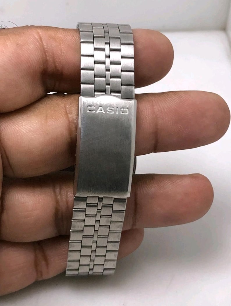May include: Close-up of a silver-toned watch band with the brand name "CASIO" engraved on the clasp. The band is made of rectangular metal links. The watch band is held by a person's hand.