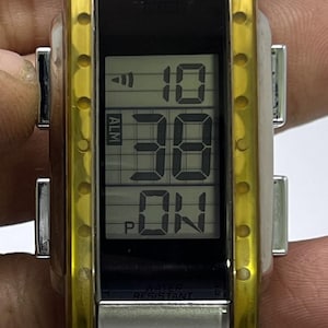 May include: A Citizen digital watch with a rectangular black display showing the time as 10:38 and the word "ON". The watch has a transparent yellow and silver case and a silver band. The watch is water resistant.