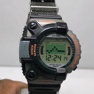 Op de afbeelding: Een zwarte en grijze Casio Cybermax digitale horloge met een groen LCD-scherm met de tijd 12:24:14. Het horloge heeft een zwarte stoffen band en de tekst "SHOCK SENSOR" en "WATER RESIST 5 BAR".