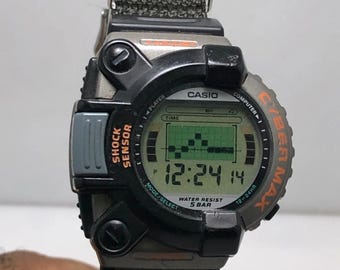 May include: A black and gray Casio Cybermax digital watch with a green LCD screen displaying the time 12:24:14. The watch has a black fabric strap and the text "SHOCK SENSOR" and "WATER RESIST 5 BAR".