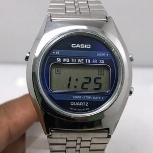 May include: A silver-toned Casio digital watch with a blue face. The watch displays the time as 1:25. The watch has a rectangular digital display and a metal band. The watch face includes the text "CASIO" and "QUARTZ".