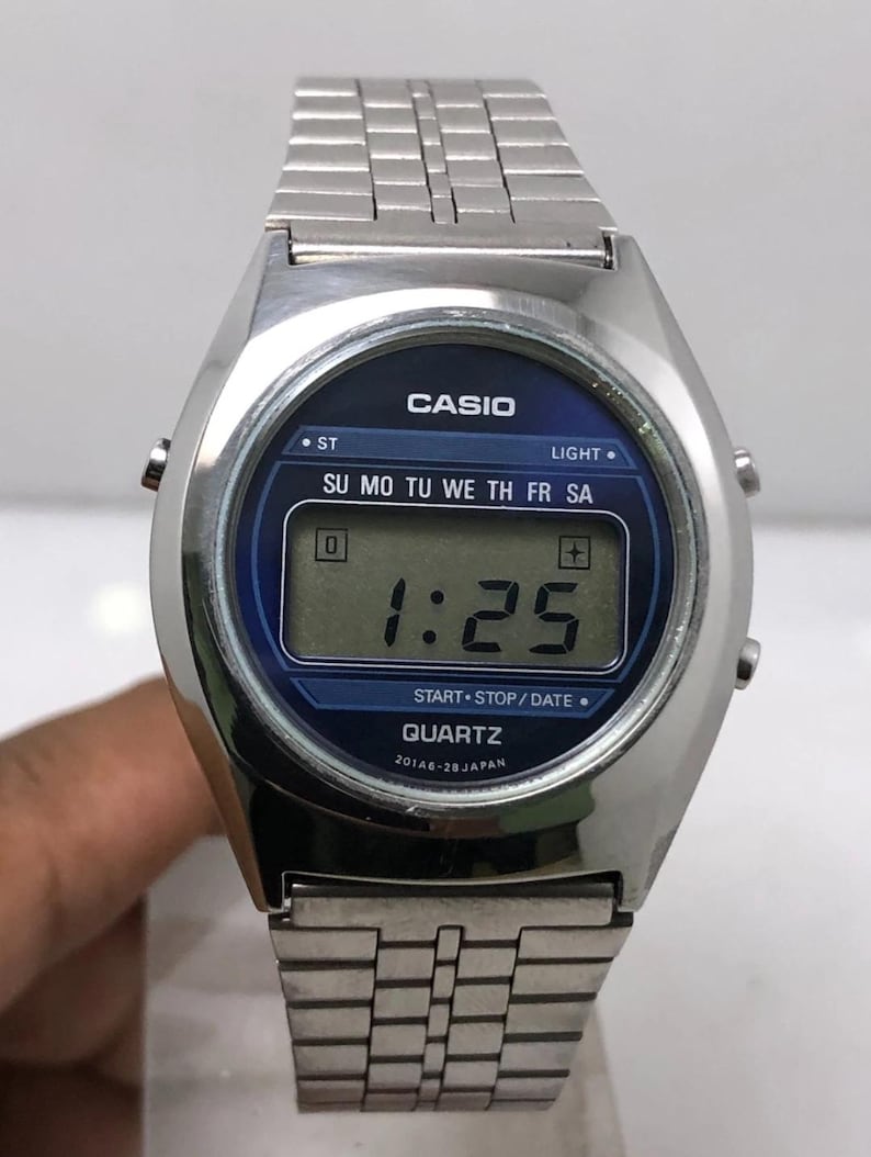 May include: A silver-toned Casio digital watch with a blue face. The watch displays the time as 1:25. The watch has a rectangular digital display and a metal band. The watch face includes the text "CASIO" and "QUARTZ".