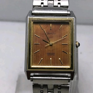 以下が含まれることがあります： 金色の文字盤が付いた長方形のシルバーの腕時計。「Chandor」と「Orient Quartz」のブランド名が表示されています。留め金が付いたシルバーのバンドが付いています。