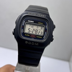 May include: Black Casio digital watch with a rectangular face. The watch displays the time, day, and date. The watch face has the text "CASIO", "WATER RESIST", and "300M". The watch is on a clear stand.