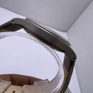 Może przedstawiać: Widok z boku srebrnego zegarka na rękę z brązowym sk&oacute;rzanym paskiem. Zegarek ma prostokątną tarczę i minimalistyczny design. Pasek jest przymocowany do koperty zegarka za pomocą małych szpilek. Zegarek jest prezentowany na przezroczystym, zakrzywionym stojaku.
