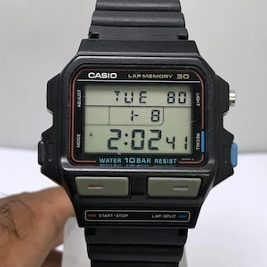 以下が含まれることがあります： 黒いカシオデジタル腕時計。長方形の文字盤と黒い樹脂バンドが付いています。文字盤には曜日、日付、時刻が表示されます。時刻、モード、ライト、スタート/ストップ、ラップ/スプリット機能を調整するためのボタンがあります。