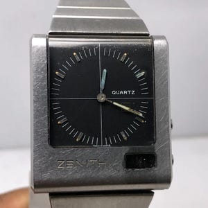 Könnte beinhalten: Eine Vintage Zenith Quarzuhr mit einem quadratischen, gebürsteten Metallgehäuse und einem schwarzen Zifferblatt. Das Zifferblatt hat weiße Markierungen und Zeiger, mit dem Wort "QUARTZ" oben aufgedruckt. Das Armband der Uhr ist ebenfalls aus gebürstetem Metall.