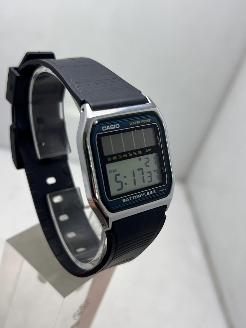 May include: A black Casio digital watch with a rectangular face. The watch displays the time as 5:17 PM and the date. The watch has a black band and a silver-colored bezel. The text "CASIO" and "WATER RESIST" are visible on the watch face.