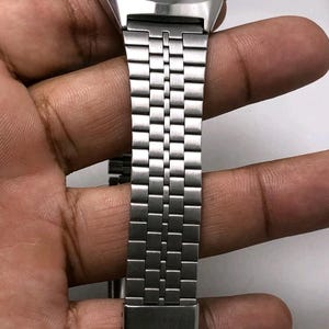 May include: Silver-toned digital watch with a rectangular face and a matching metal band. The watch face has a blue background with white text. The band is made of small, rectangular metal links.