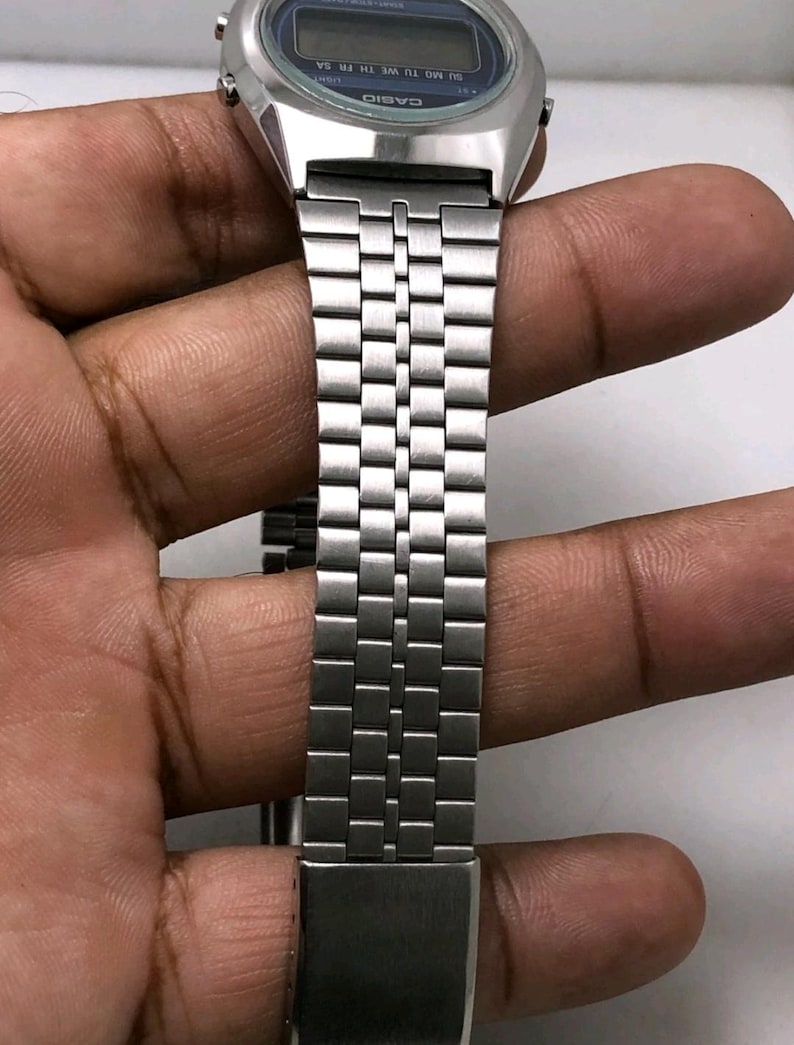 May include: Silver-toned digital watch with a rectangular face and a matching metal band. The watch face has a blue background with white text. The band is made of small, rectangular metal links.