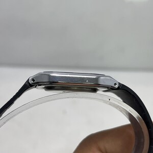 May include: A silver rectangular wristwatch with a black leather band. The watch face is silver and the band is attached to the watch case with small silver pins. The watch is displayed on a clear, curved stand.
