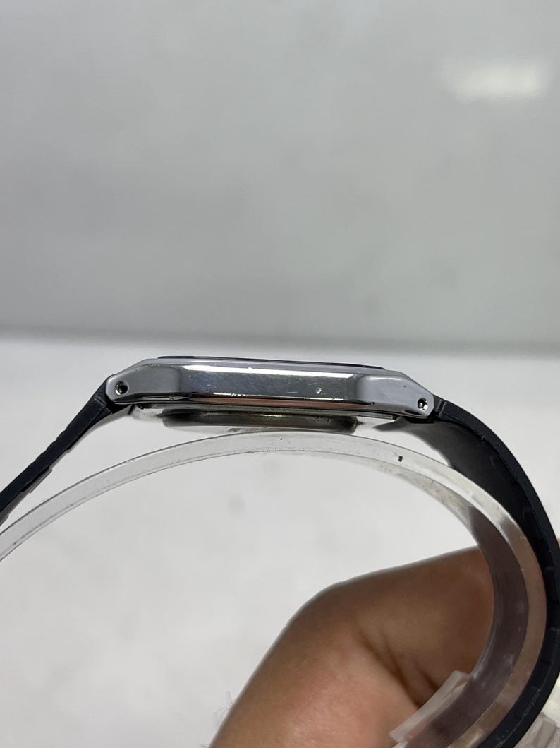 May include: A silver rectangular wristwatch with a black leather band. The watch face is silver and the band is attached to the watch case with small silver pins. The watch is displayed on a clear, curved stand.
