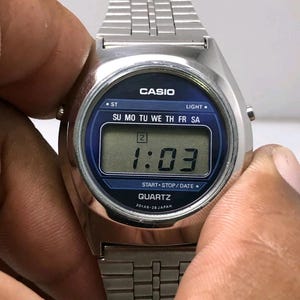 May include: A silver Casio digital watch with a blue face displaying the time as 1:03. The watch has a rectangular digital display and a silver metal band. The watch face includes the text "CASIO", "QUARTZ", and the days of the week.