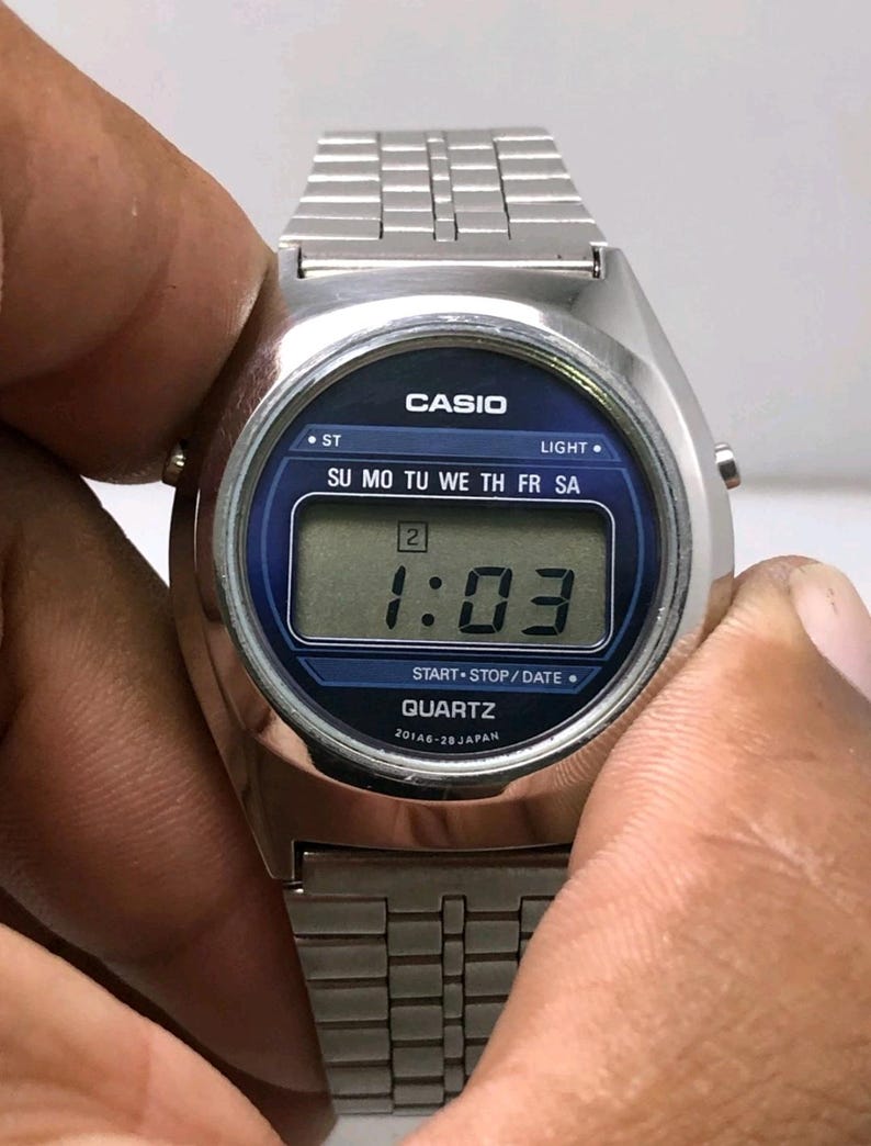 May include: A silver Casio digital watch with a blue face displaying the time as 1:03. The watch has a rectangular digital display and a silver metal band. The watch face includes the text "CASIO", "QUARTZ", and the days of the week.