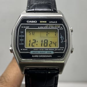 May include: A silver Casio digital watch with a black leather band. The watch face displays the time, date, and various functions. The watch is water-resistant to 100 meters.