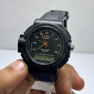 Könnte beinhalten: Schwarze Casio-Uhr mit rundem Zifferblatt, orangefarbenem Sekundenzeiger und Digitalanzeige. Die Uhr hat ein schwarzes Kautschukarmband und mehrere Knöpfe zur Einstellung der Einstellungen. Auf dem Zifferblatt steht "CASIO" und "TI-DEPTH METER".
