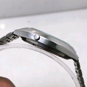 May include: A silver-toned wristwatch with a rectangular face and a linked metal band. The watch has a light-colored face and a small button on the side. The watch is displayed on a clear, curved stand.