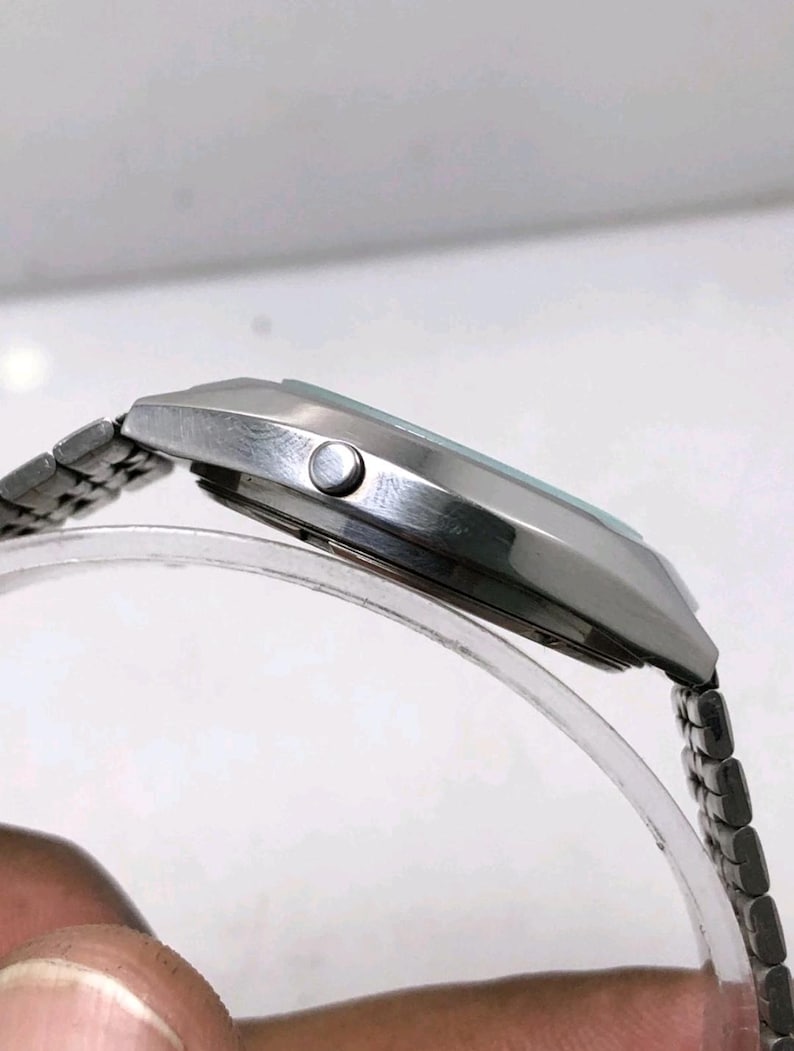 May include: A silver-toned wristwatch with a rectangular face and a linked metal band. The watch has a light-colored face and a small button on the side. The watch is displayed on a clear, curved stand.