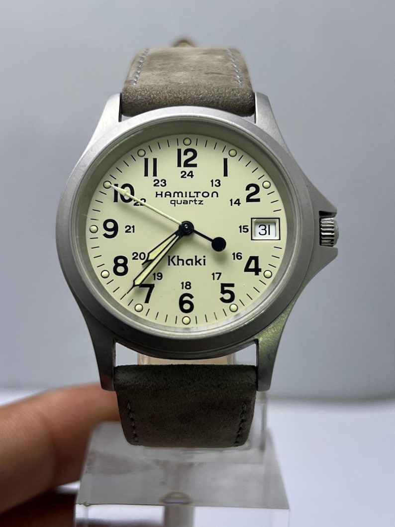 Może przedstawiać: Srebrny zegarek kwarcowy Hamilton Khaki z kremową tarczą i szarym zamszowym paskiem. Tarcza zegarka wyświetla czarne cyfry, okienko daty i nazwę marki. Zegarek ma klasyczny, użytkowy design.
