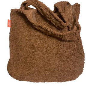May include: A brown tote bag made of textured, fuzzy sherpa fleece material. The bag features two matching shoulder straps and a small, rectangular orange fabric tag attached to the side seam. The overall design is simple, soft, and plush.