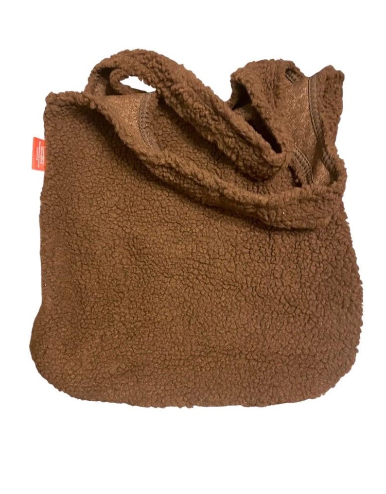 May include: A brown tote bag made of textured, fuzzy sherpa fleece material. The bag features two matching shoulder straps and a small, rectangular orange fabric tag attached to the side seam. The overall design is simple, soft, and plush.