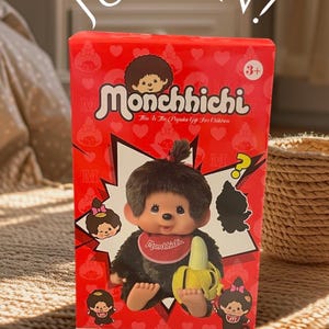 May include: Red box featuring a Monchhichi doll with a banana. The box has the text "JUST IN!" and "Monchhichi" with the age range "3+". The doll is black with a red bib and a yellow banana.