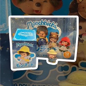 Puede incluir: Un juego de cajas de muñecas Monchhichi, con varias figuras pequeñas con diferentes atuendos. La caja es azul y blanca, con el logotipo de Monchhichi y texto japonés. El juego incluye 12 piezas.
