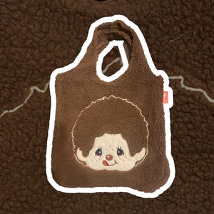 May include: A brown sherpa fleece tote bag featuring an embroidered character face with a round brown hairstyle, a light beige complexion, and a small red tongue sticking out. The bag has two integrated handles and a small red side tag.