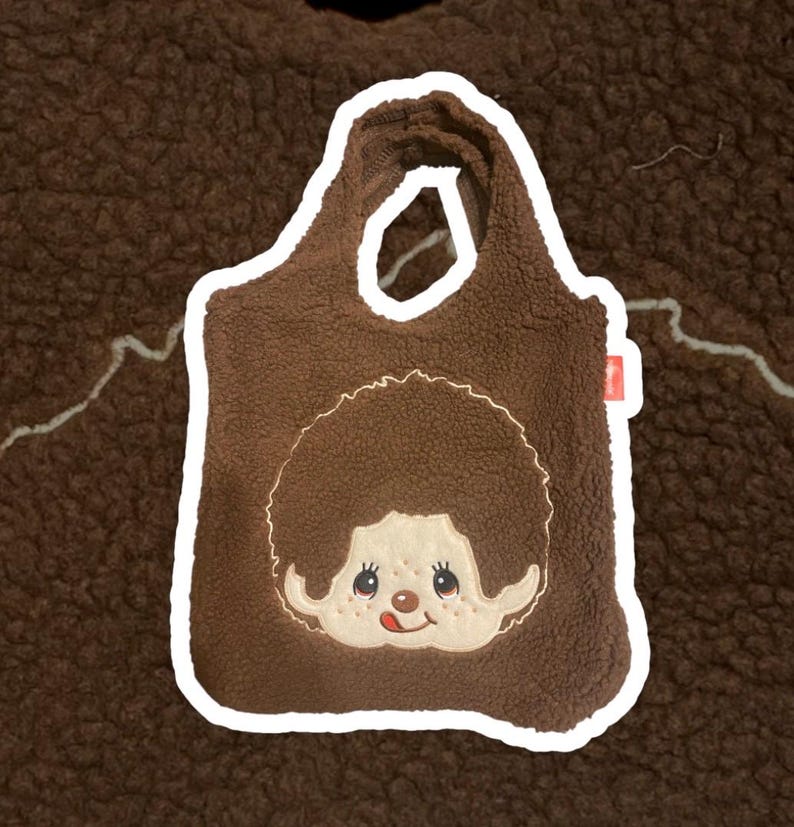 May include: A brown sherpa fleece tote bag featuring an embroidered character face with a round brown hairstyle, a light beige complexion, and a small red tongue sticking out. The bag has two integrated handles and a small red side tag.