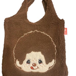 May include: A brown sherpa fleece tote bag featuring a large embroidered applique of a character with a round face, freckles, and a tongue sticking out. The bag has two integrated handles and a small red rectangular tag on the right side.