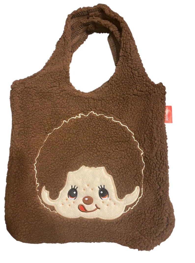 May include: A brown sherpa fleece tote bag featuring a large embroidered applique of a character with a round face, freckles, and a tongue sticking out. The bag has two integrated handles and a small red rectangular tag on the right side.