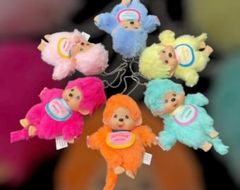 May include: A collection of six plush monkey dolls arranged in a circle, each featuring a different vibrant fur color including blue, yellow, turquoise, orange, hot pink, and light pink. Each doll has a plastic face and a small bib with text.