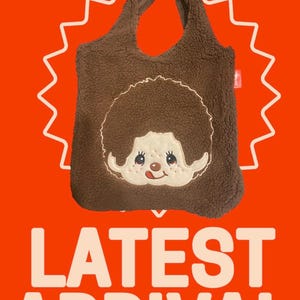 May include: A brown fuzzy tote bag featuring a cream-colored embroidered face of a character with a large round hairstyle, freckles, and a small tongue sticking out. The bag has two integrated handles and a soft, textured fleece-like material.