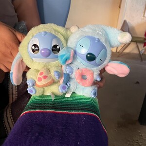May include: Two blue plush toys resembling a cartoon character, each wearing a hooded outfit. One is light green with a pink and yellow pastry, the other is light blue with a pink donut. Both have large eyes and are holding treats.