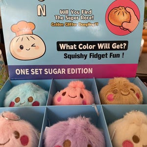 Puede incluir: Una caja expositora con seis llaveros de peluche con forma de bola de masa en varios colores pastel, como azul, rosa, beige, lavanda y crema. Cada peluche tiene una cara sonriente con ojos bordados y mejillas rosas. La caja incluye el texto: Rainbow Mystery, Squishy Bun y One Set Sugar Edition.
